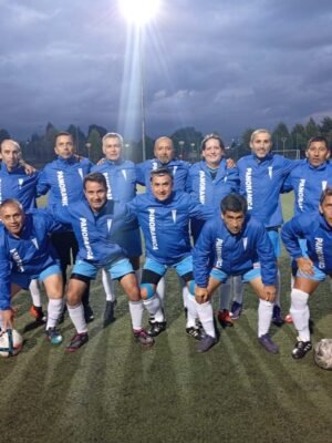 Equipo de fútbol adulto posa en cancha sintética bajo iluminación nocturna. Todos lucen chaquetas personalizadas color azul con el nombre “PANORÁMICA”, destacando la confección profesional de vestuario deportivo Kotoduo para adultos.
