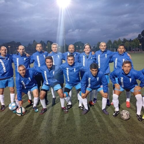 Equipo de fútbol adulto posa en cancha sintética bajo iluminación nocturna. Todos lucen chaquetas personalizadas color azul con el nombre “PANORÁMICA”, destacando la confección profesional de vestuario deportivo Kotoduo para adultos.